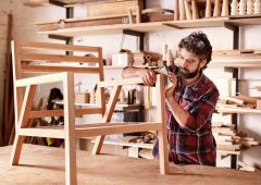 Serious furniture designer carefully sanding a chair frame that he is busy manufacturing in his woodwork studio, with shelves of wooden items behind him